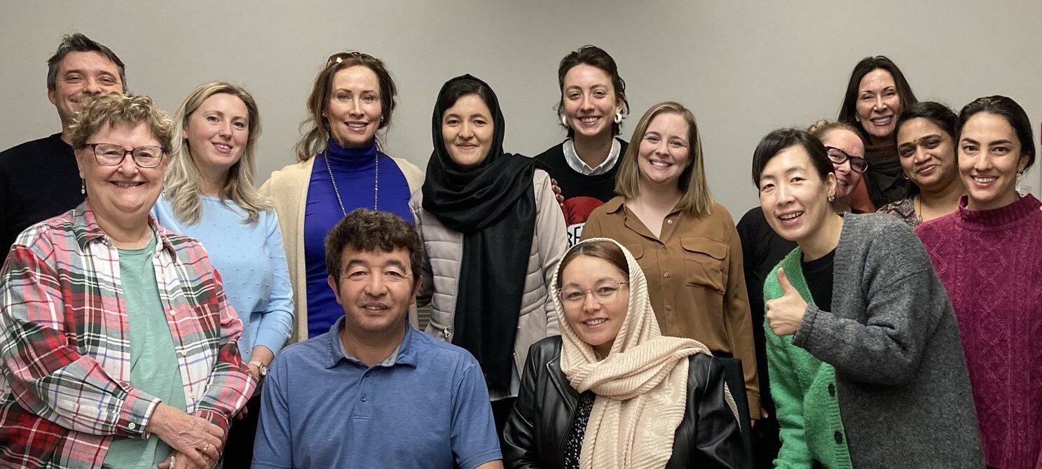 A diverse group of adults posing and smiling together in a classroom setting, with some sitting and others standing behind them.