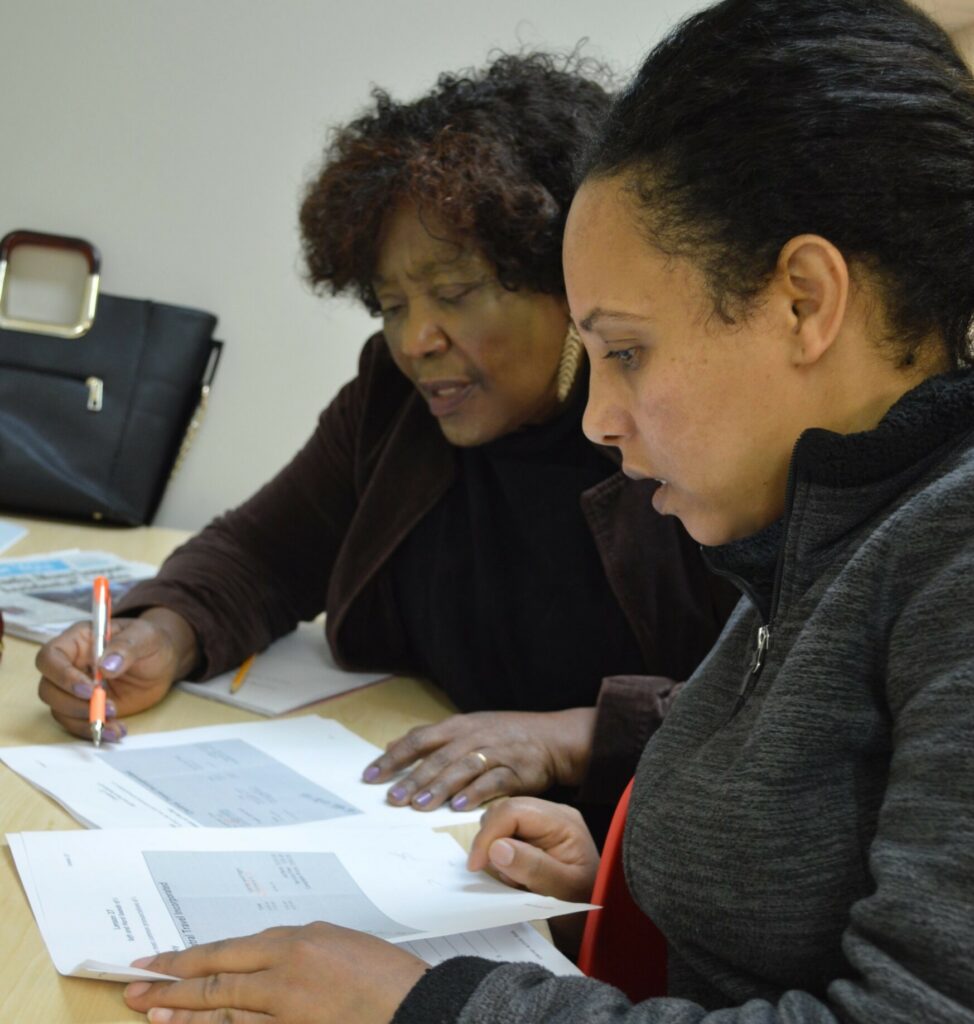 Two women sitting at a table working on documents together, one holding a pen and the other closely reviewing the papers.